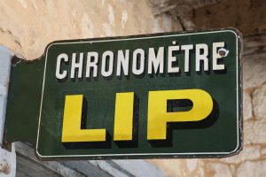 Väggskylt "CHRONOMÈTRE LIP" i grönt och gult, monterad på en ljus vägg.